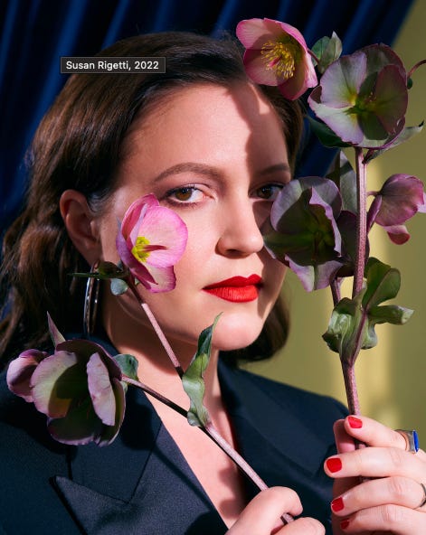 A photograph of Susan with red lipstick and a black jacket, holding pink flowers up in front of her face.