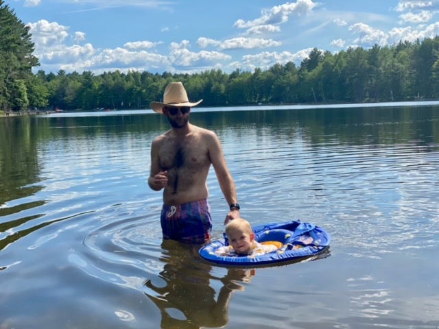 Man in cowboy hat stands in thigh-high water while child in floatation device floats.