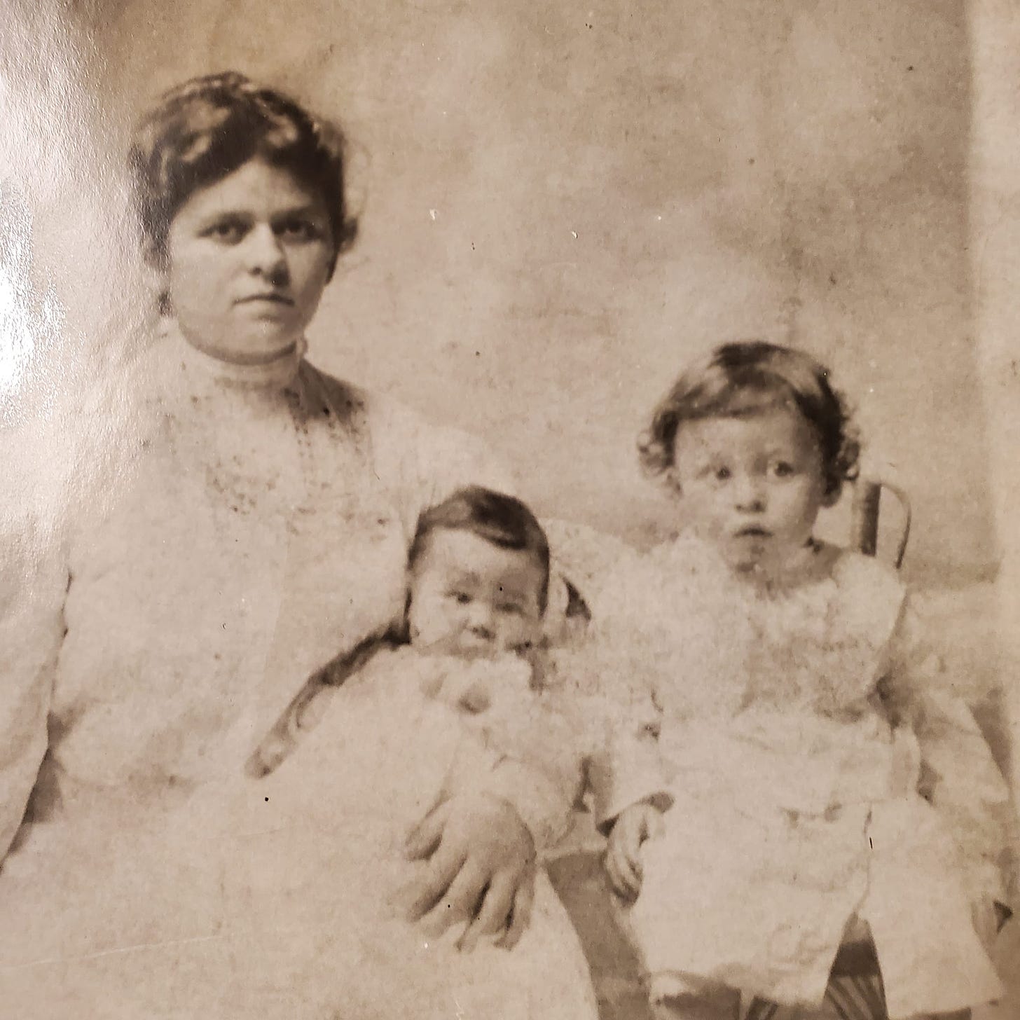 A black and white photo of a dark-haired white woman, an infant, and a toddler, all dressed in white dresses.