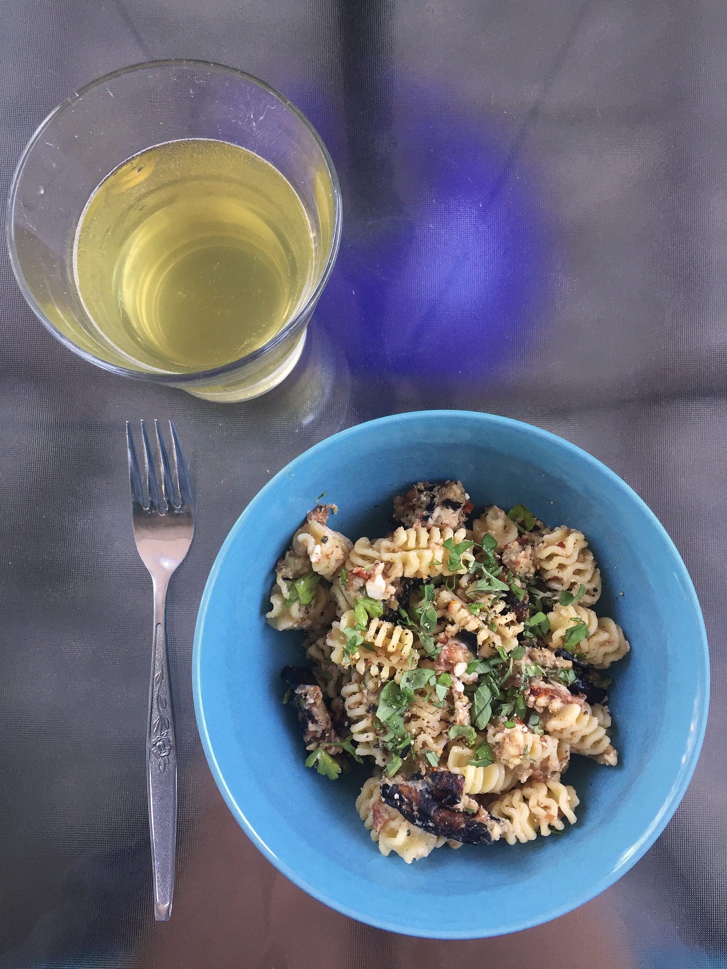 On an outdoor table, a blue bowl of pasta salad with radiatore and charred eggplant. Pieces of feta and lots of green herbs are visible throughout, along with small pieces of sun-dried tomato. Next to it is a glass of pale saison.