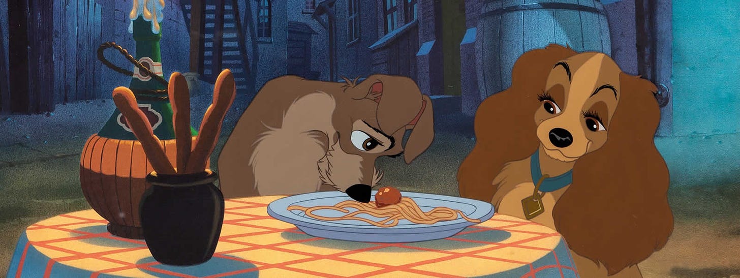 One cartoon dog noses a meatball toward another during a romantic spaghetti dinner.
