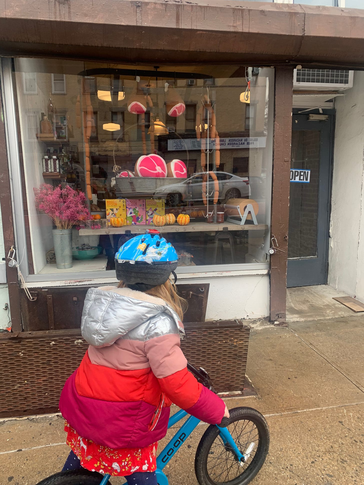 girl on bicycle in puffy coat stops in front of ham store, with toy hams in window