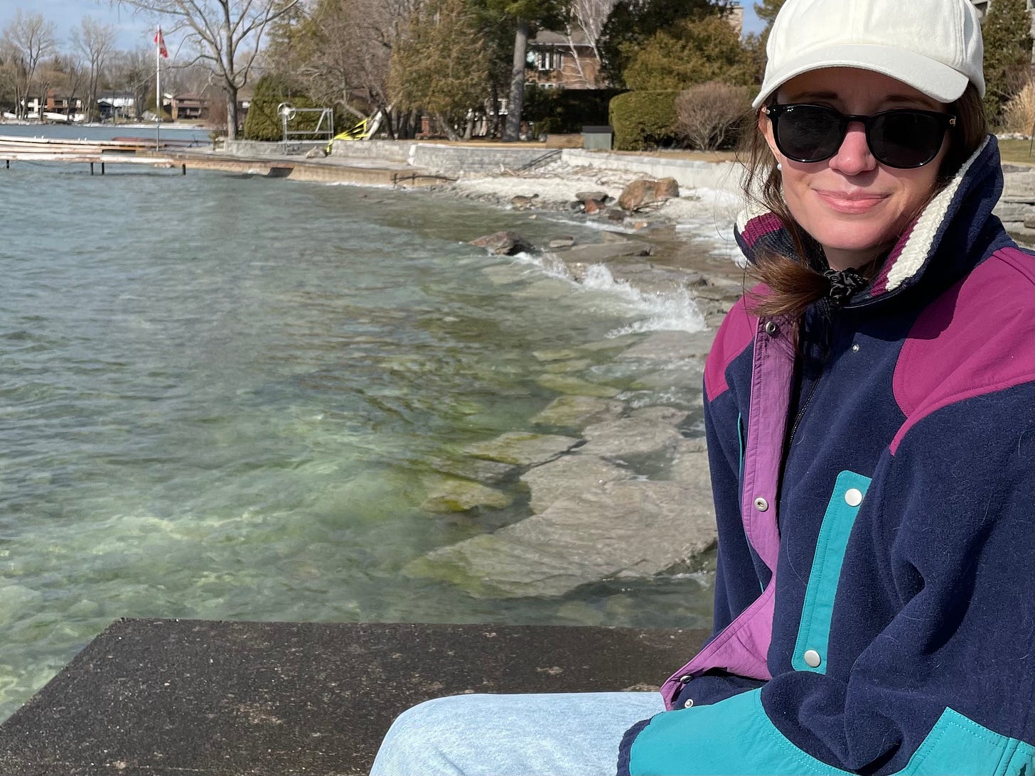 a white woman sits by a lake in a colorful fleece jacket, sunglasses, and a white hat