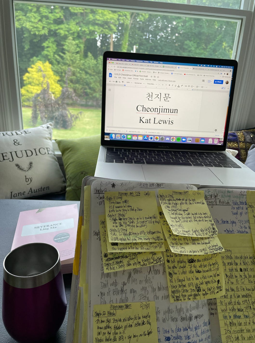 A photo of Kat's writing desk. Her laptop is opened to her novel manuscript titled, CheonJiMun. There is a composition notebook opened to a page of novel notes with many post-it notes stuck to it. The novel, Severance by Ling Ma, is also visible on the desk.
