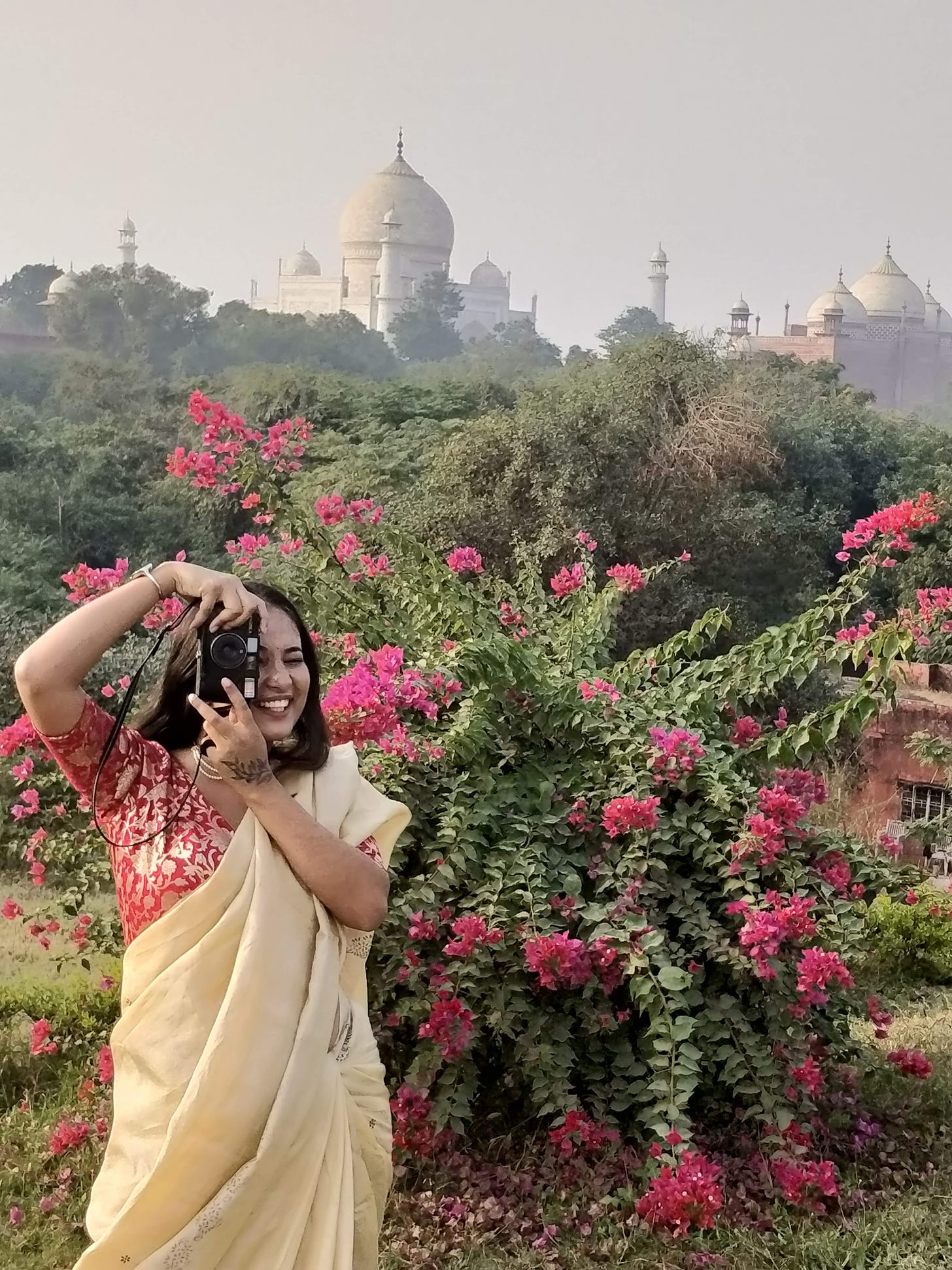 a woman in a beige saree, taking a picture of the photographer with an old film camera, in front of a bougainvillea. in the distance the taj mahal is visible