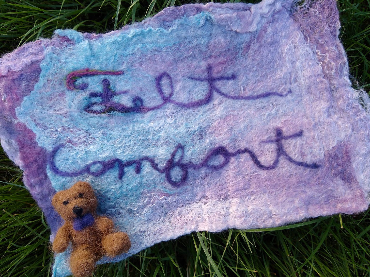Small needle-felted teddy bear on a purple and blue piece of felt that says Felt Comfort in cursive, on top of some grass