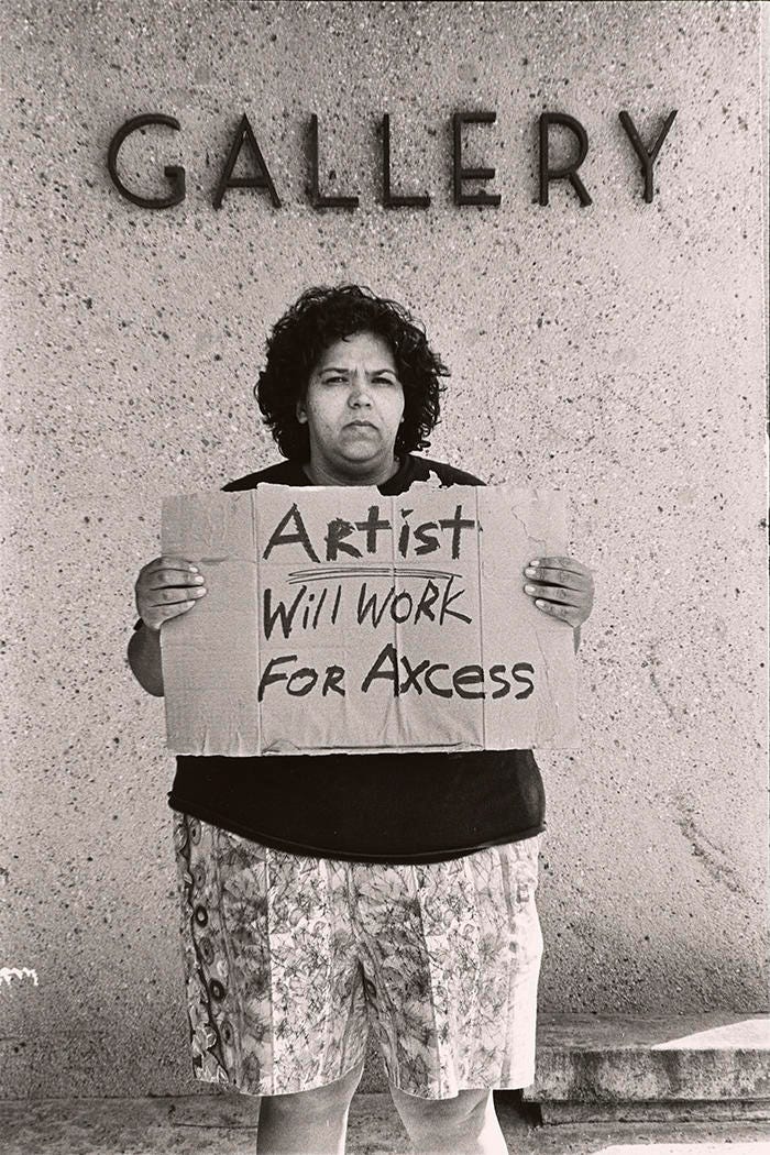 The artist stands outside a building marked “GALLERY,” holding a cardboard sign that says “Artist Will Work for Axcess.”