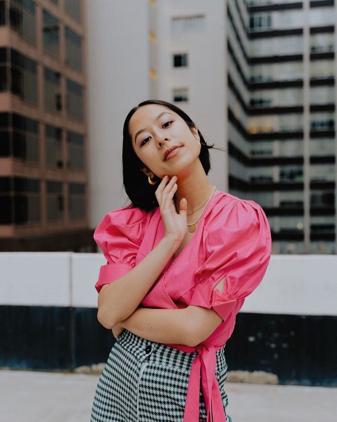 A young Asian Australian woman, standing on a rooftop in front of city buildings, posing with her hand on her chin and wearing a bright pink blouse