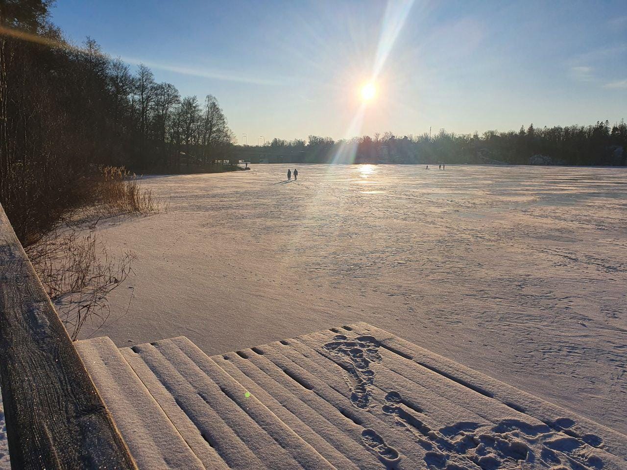 Um lago imenso coberto de neve, onde ao longe pessoas caminham e o sol brilha.