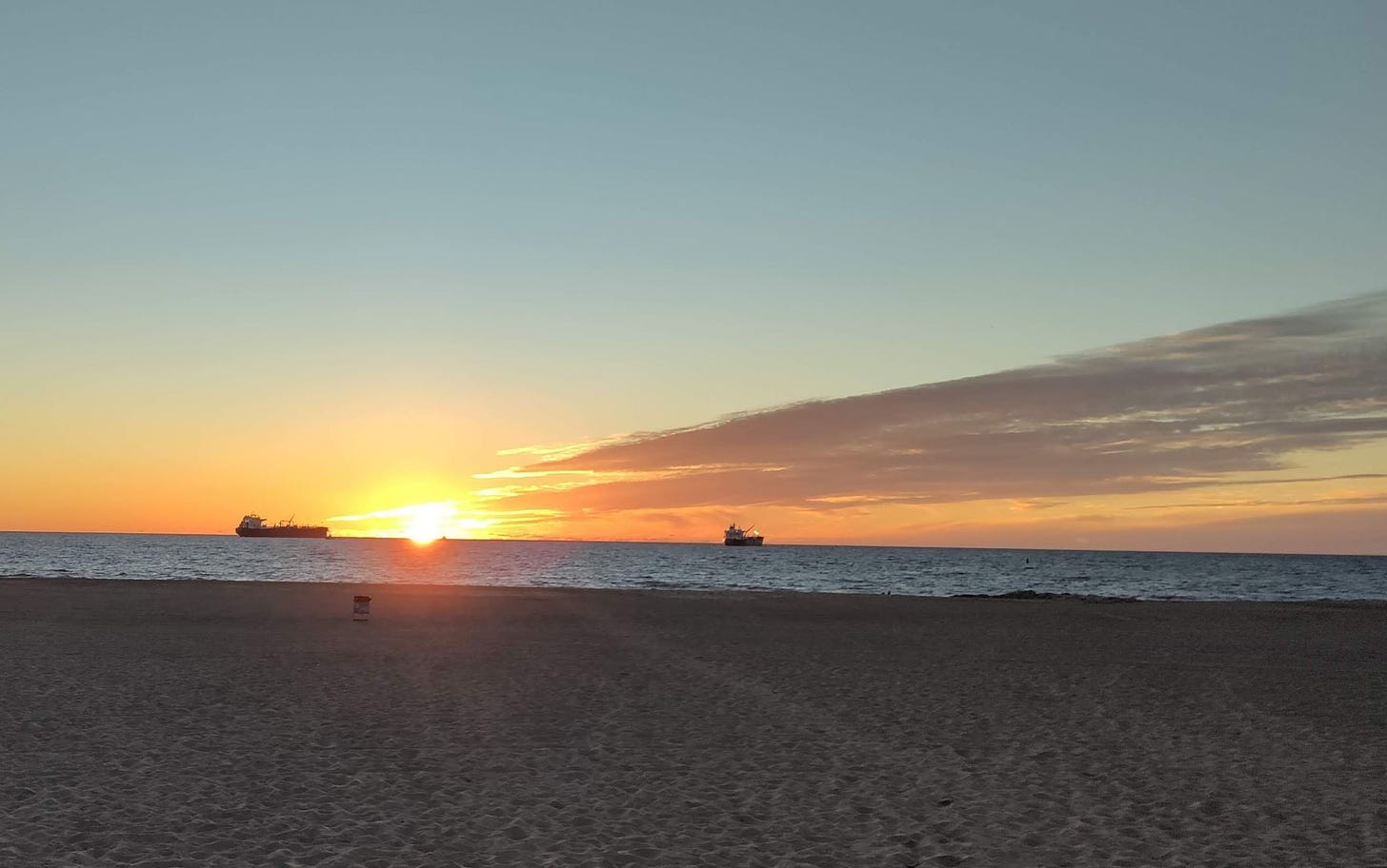 sun setting on the Pacific Ocean with two cargo ships in the background and beach in the foreground
