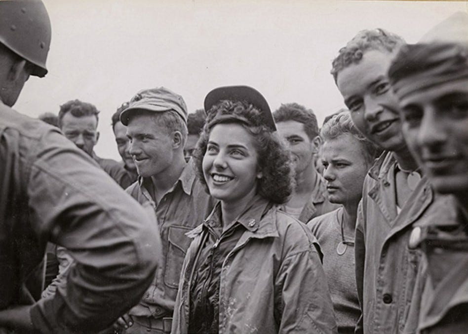 A photo of Jane Kendeigh in the midst of a group of soldiers and/or Marines. She is a pretty woman with curly hair. She is smiling up at someone (maybe an officer?) who is standing in front of the crowd.