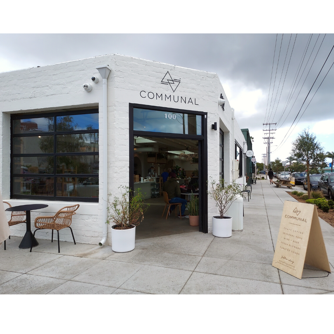 The front of a coffee shop. A corner view of a white concrete block building with an open door with a black frame. The intertwined triangle logo with the name "Communal" is painted above the door fram. You can see the sidewalk along the right side of the fram with a picnic table in the distance under telephone wires and next to parked cars on the street. A raw wood a-fram sign welcomes you into the cafe and two large potted plants frame the doorway.