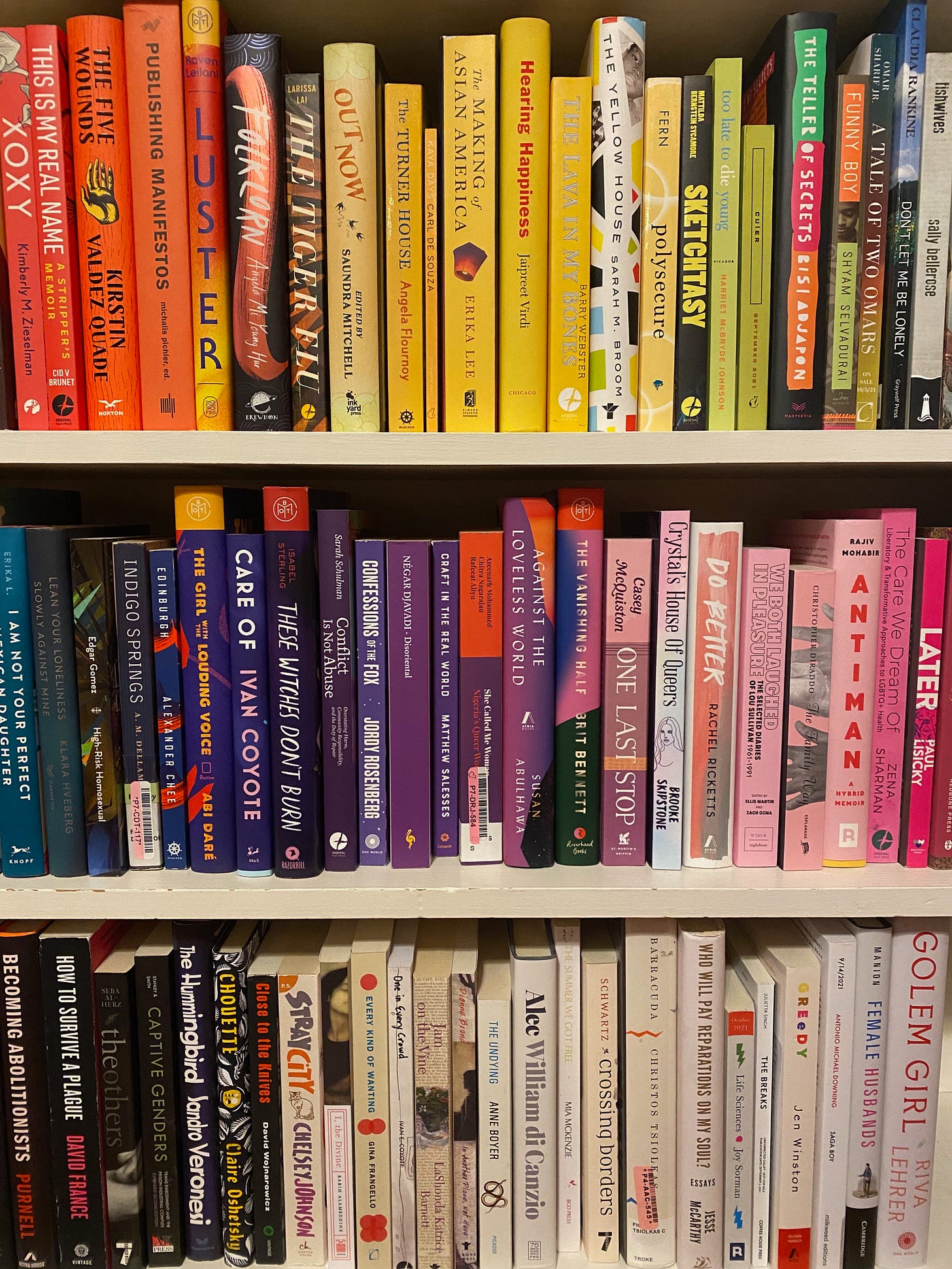 Three shelves of books, the spines arranged in the colors of a rainbow from top to bottom.