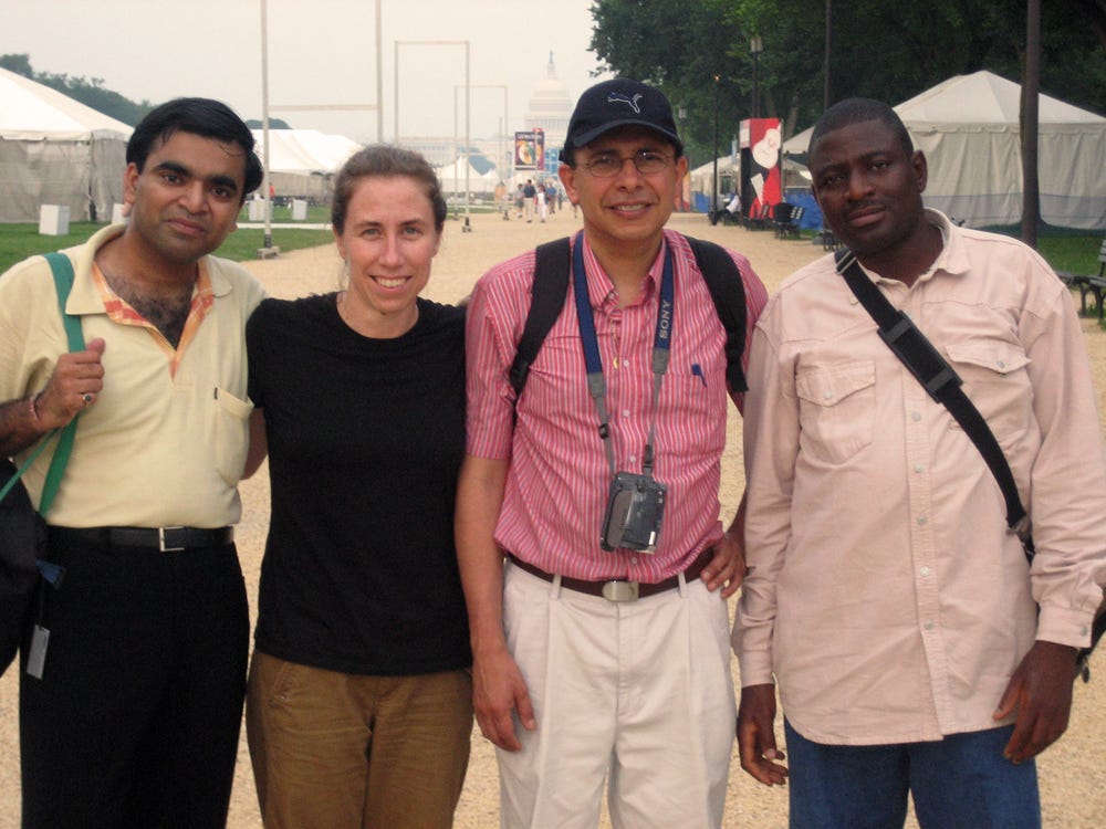 Four people stand lookng at the camera, with the Capitol building in the far distance. They are a South Asian man in a yellow shirt, a white person in a black shirt, a Latin American man in a red striped shirt and black hat, and a Nigerian man in a white button-down shirt.