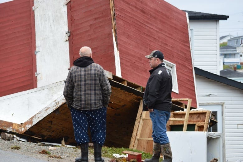 Two men wearing rubber boots in Port aux Basques survey the devastation of their town after hurricane Fiona. Near them, a shed that was lifted by the waves has landed upside down on a neighbouring lawn.