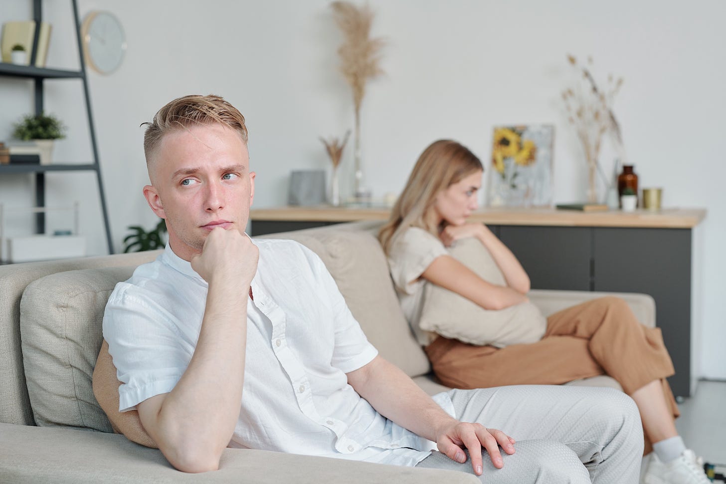 A couple with concerned faces sit apart from one another on the couch.