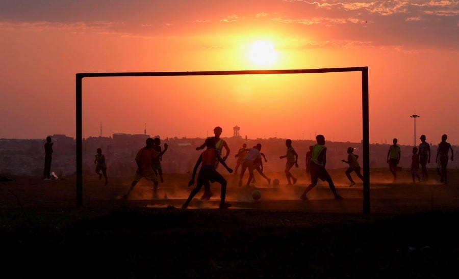 A soccer game takes place at sunset on a dusty pitch.