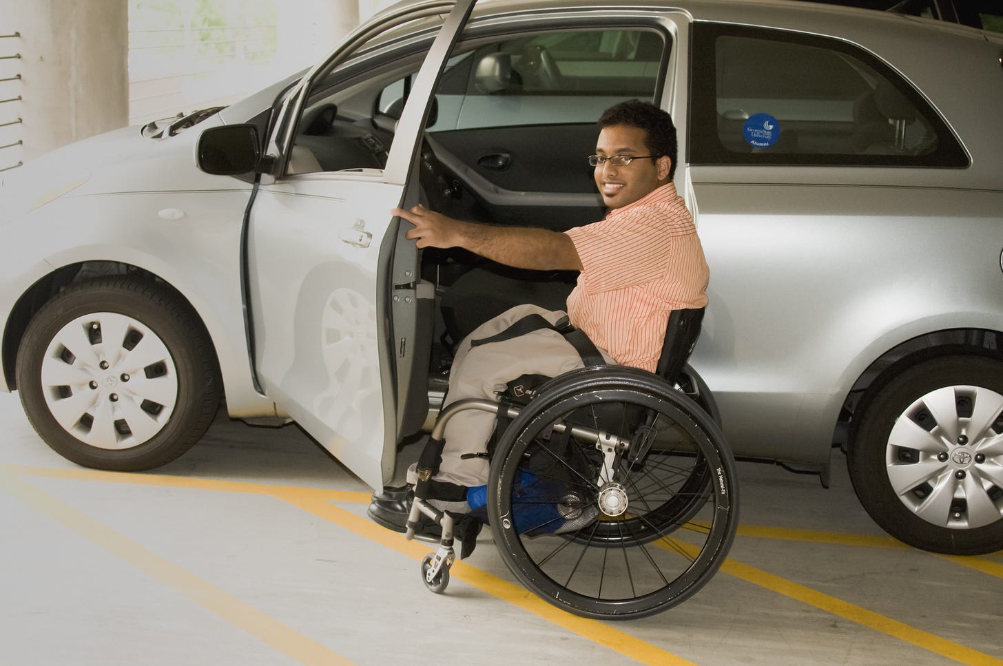 a Black man wheelchair user is wearing an orange and white striped shirt and tan trousers. He is facing the camera and grinning as he holds a car door open. The car is silver and parked next to yellow lines.