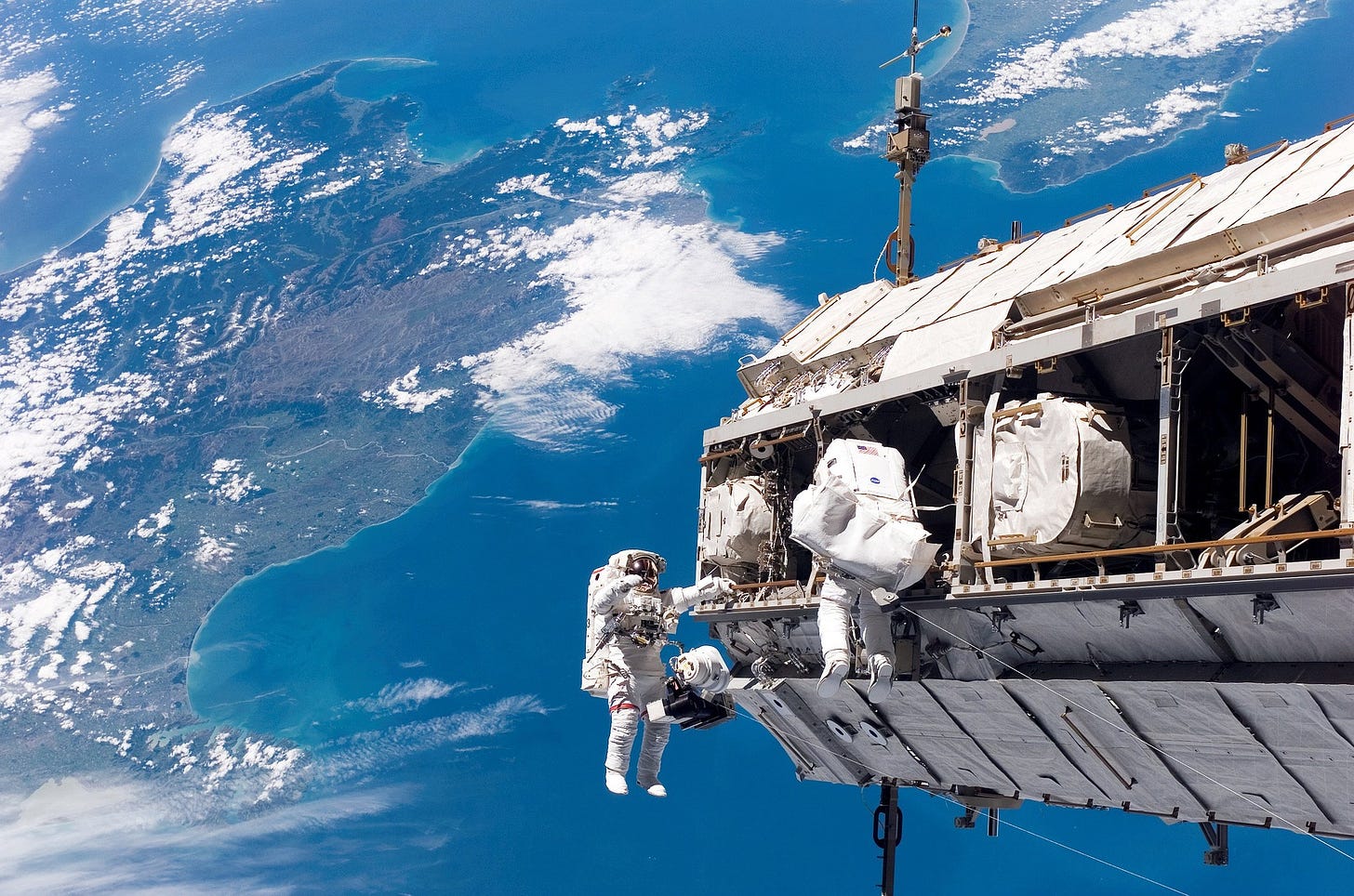 picture of two astronauts on a spacewalk on the International Space Station. in the background is the surface of the Earth, over the ocean and a large island