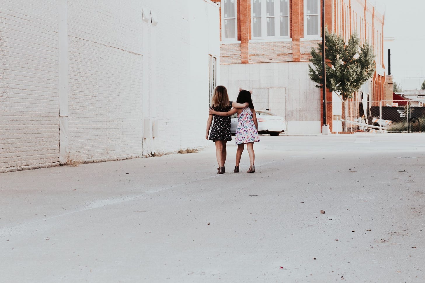 Two little girls in cute dresses walk arm and arm down a city alley.