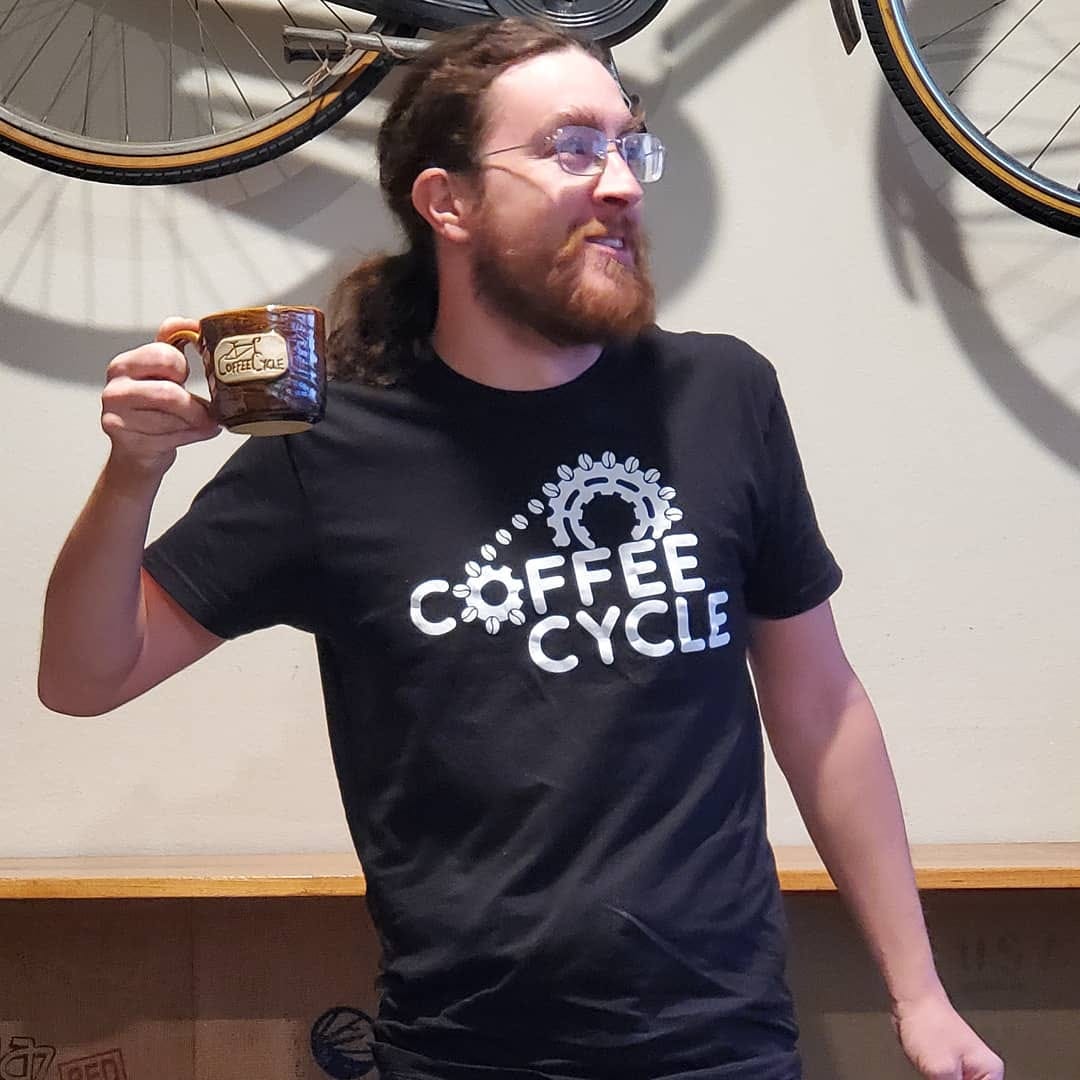 Chris O'Brien, founder of Coffee Cycle coffee shop holding a cup of coffee. A bicycle hangs on the wall behind him.