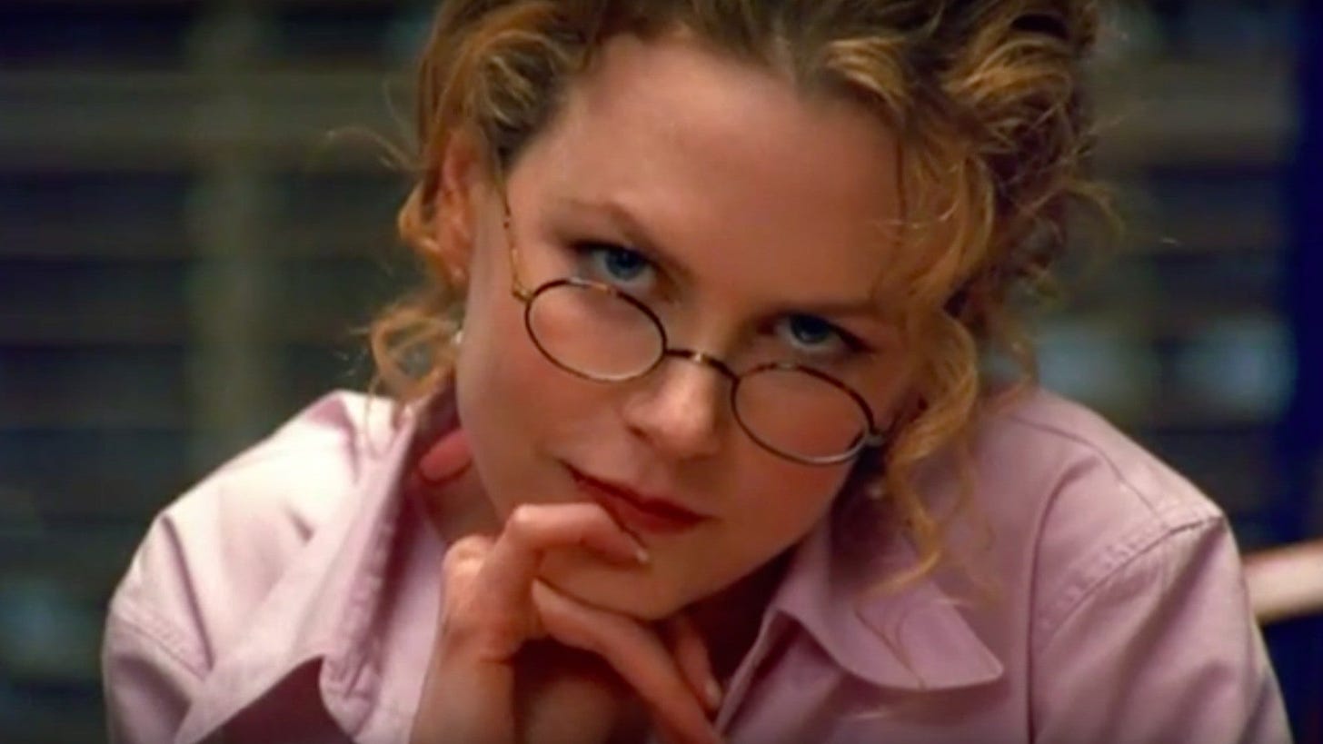 Film still from Eyes Wide Shut. Nicole Kidman stares into the camera with her hand on her chin, wearing round glasses and a dress shirt.