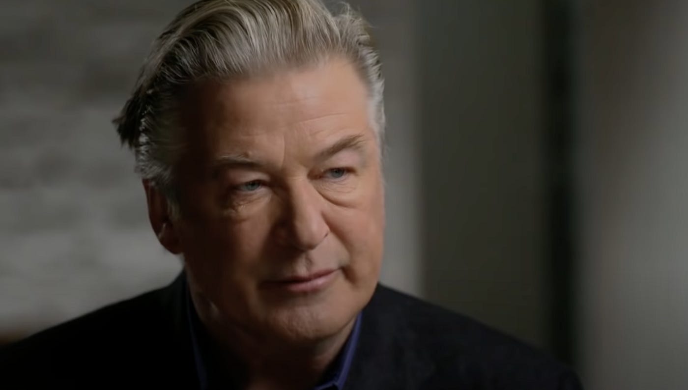 Image description: Alec Baldwin is wearing a black suit with a dark blue shirt, his grey hair combed back and with a serious expression on his face. Behind him is a blurred grey background.