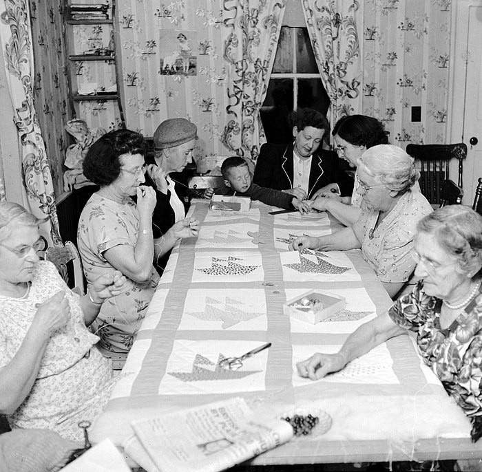 1950's Quilting Bee. Seven women sit around sewing a quilt in-progress, while a child reaches in to grab something.