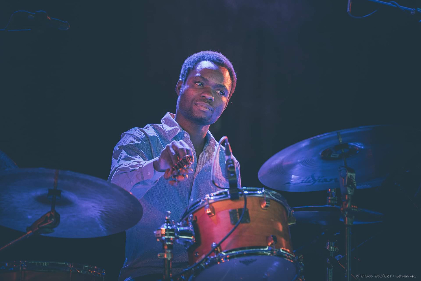 A man sits at a drum set holding an instrument with small bells in his hand. He appears to be listening to the music.