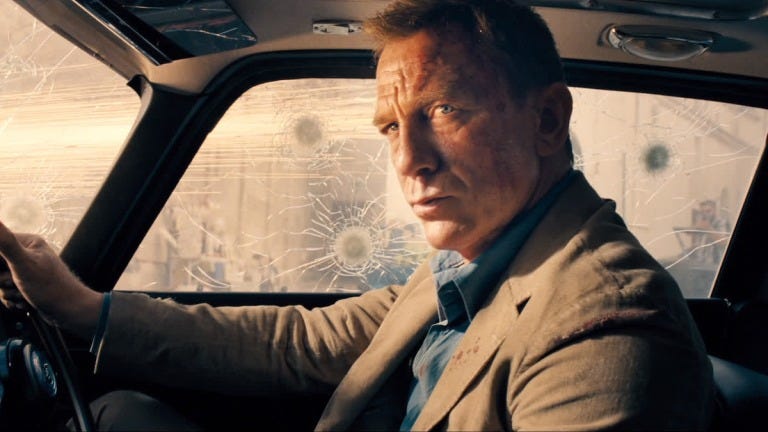James Bond (played by Daniel Craig) looks witheringly at camera while sitting inside his Astin Martin DB5, which is under attack by unknown gunmen, in this still image from No Time to Die.