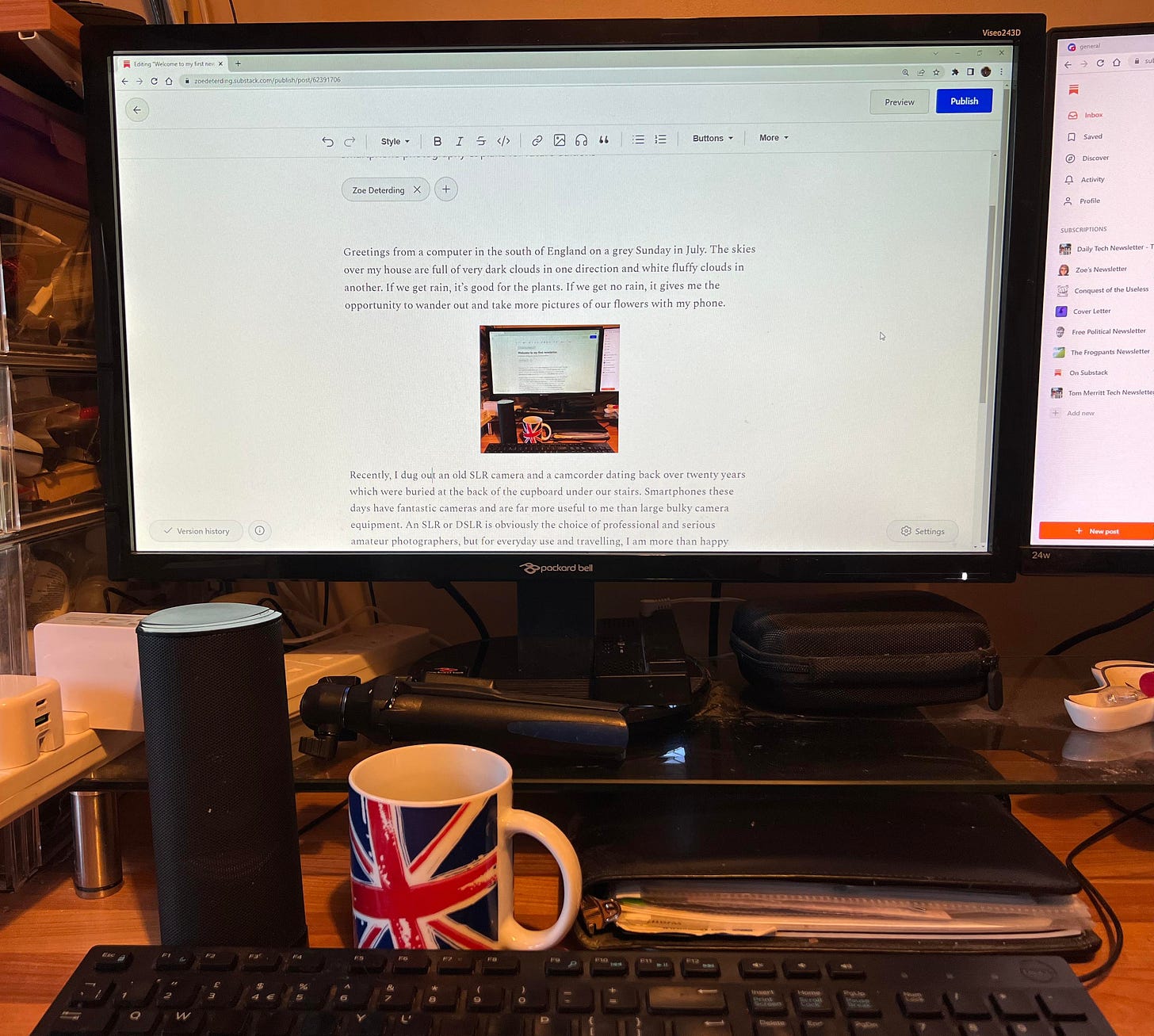 photograph of my physical desktop area, a computer screen, a mug of coffee with the UK flag on it, part of a computer keyboard and one computer speaker to one side, part of another computer screen to the right.