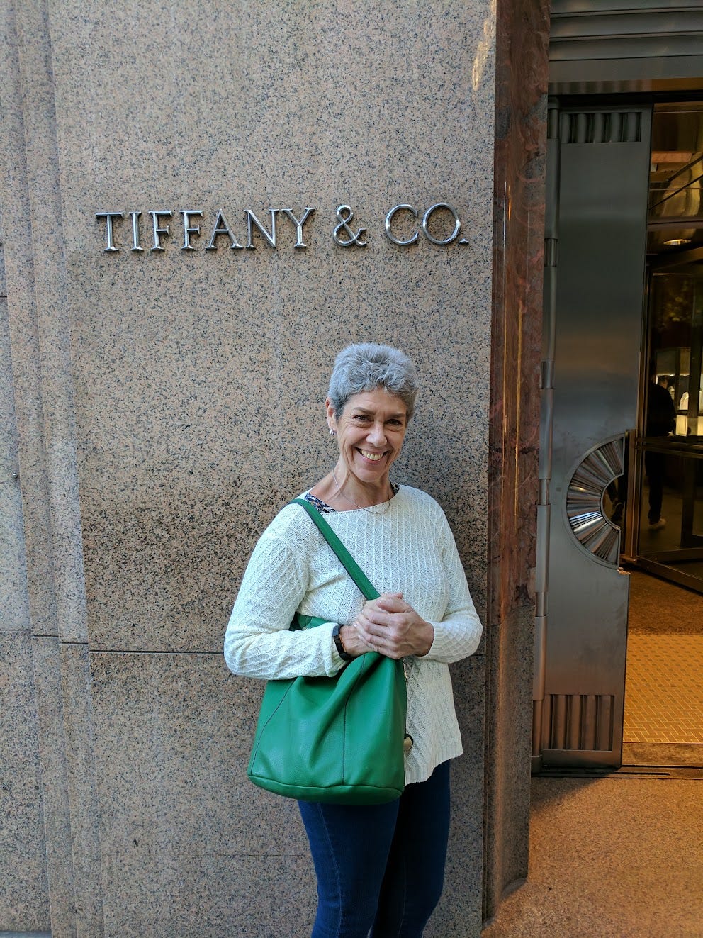 A white woman with white hair holds a green purse in front of a Tiffany sign