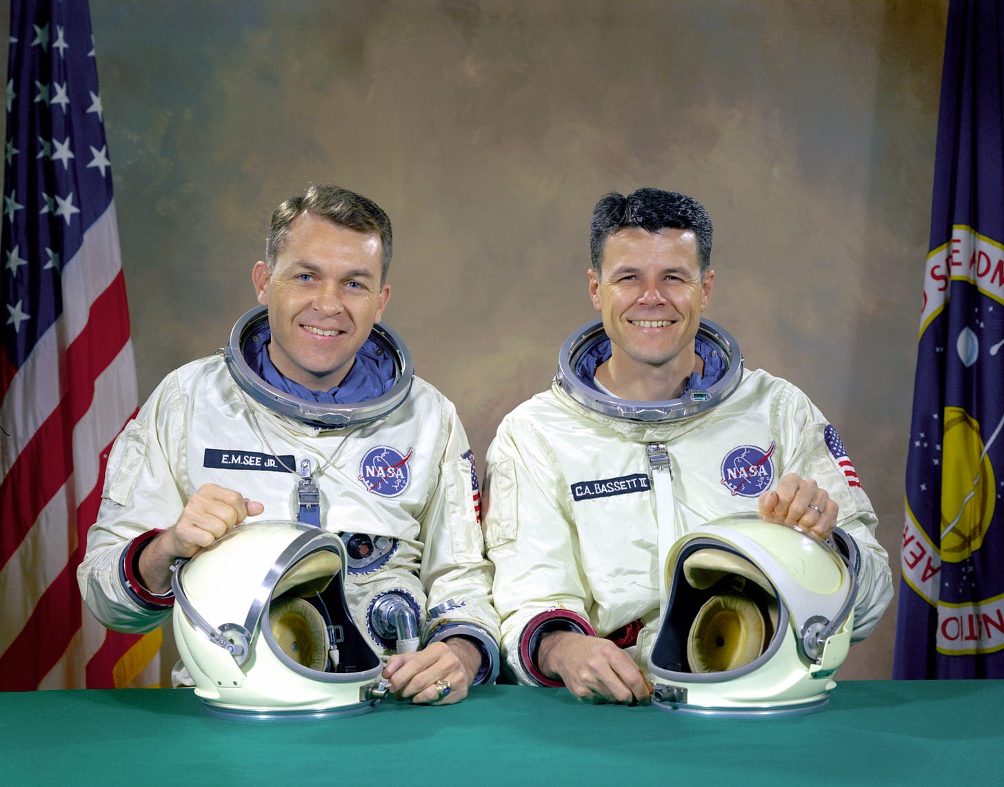 Official NASA photo of Elliot See and Charles Bassett, in spacesuits, but with their helmets on a table in front of them.  An American flag and a NASA flag provide a backdrop.