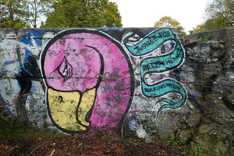 A wall with graffiti of a large pink bird's head, bowed and crying. On the right is a painted banner saying 'Justice for Belly Mujinga, Black Lives Matter'.