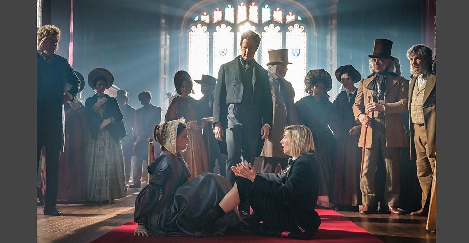 This image shows the Thirteenth Doctor (played by Jodie Whittaker) and polymath Ada Lovelace (played by Sylvie Briggs) sitting on the floor of London's Royal Gallery of Practical Science in the year 1834. The inventor Charles Babbage (played by Mark Dexter) stands behind them, in the center, amidst several attendees looking at the seated Doctor & Ada.