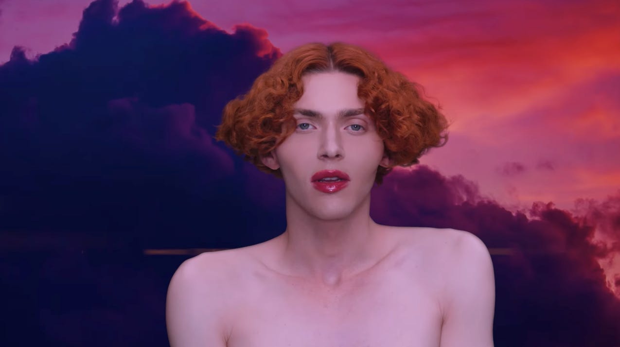 image of the artist SOPHIE from the video for "it's okay to cry," facing the camera with bare shoulders and bright red lipstick that matches the cloudy sky in the background 