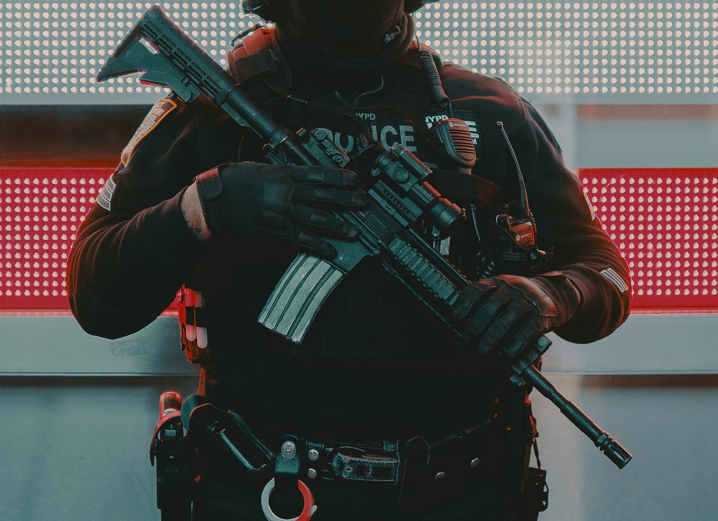Cropped photo of a heavily armed police officer in front of a neon red and white background.