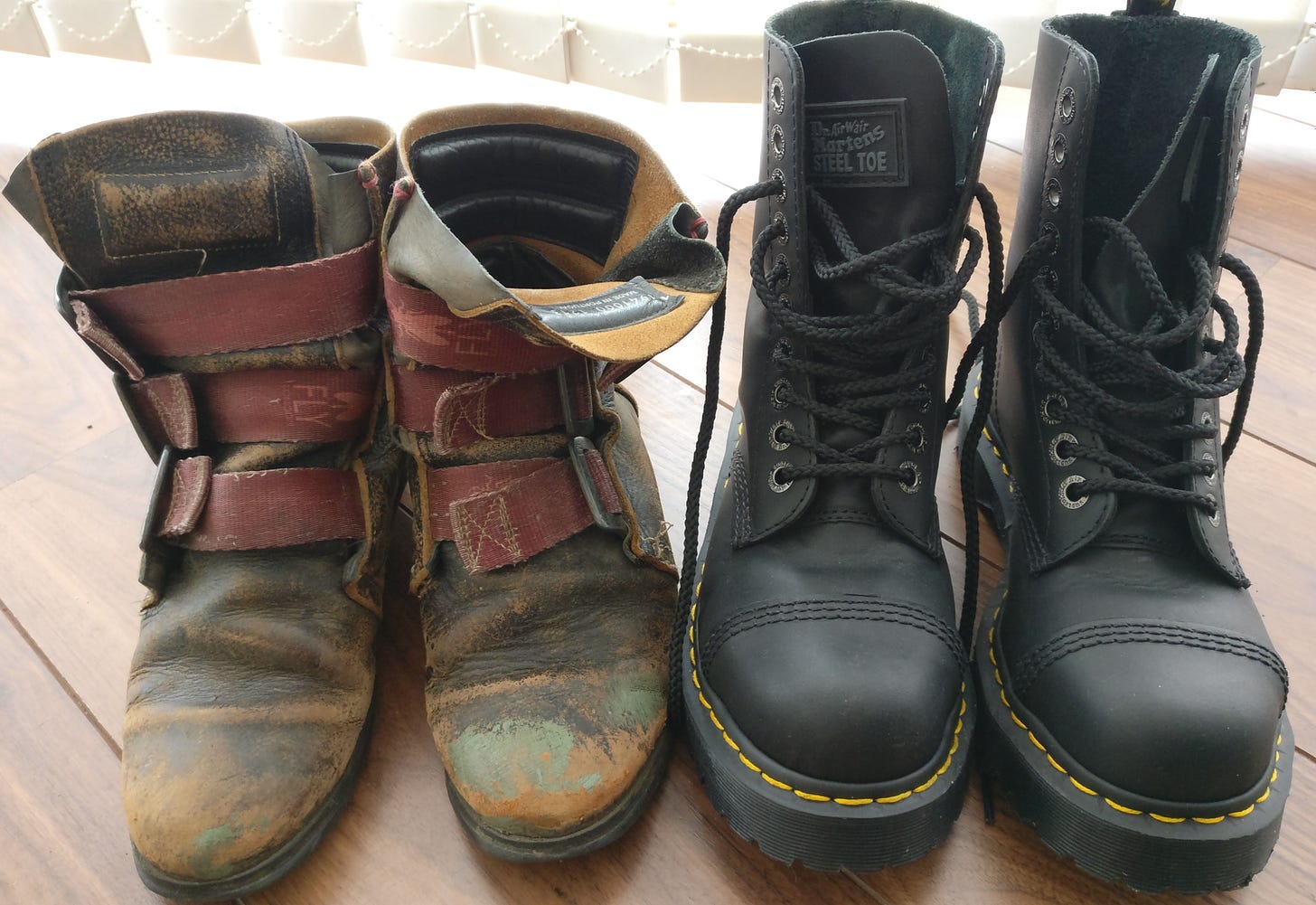 image description: a photo of two pairs of boots on a slatted wood-effect floor, with white vertical blinds hanging on the window behind them. on the left are a much-loved, totally knackered old pair of brown leather fly london pirate-style boots with three seatbelt-canvas straps. on the right are a brand new pair of black leather, steel toecapped DocMartin boots (a generous gift from a kind friend a few years ago, after seeing the state of the other ones, with their soles hanging off).