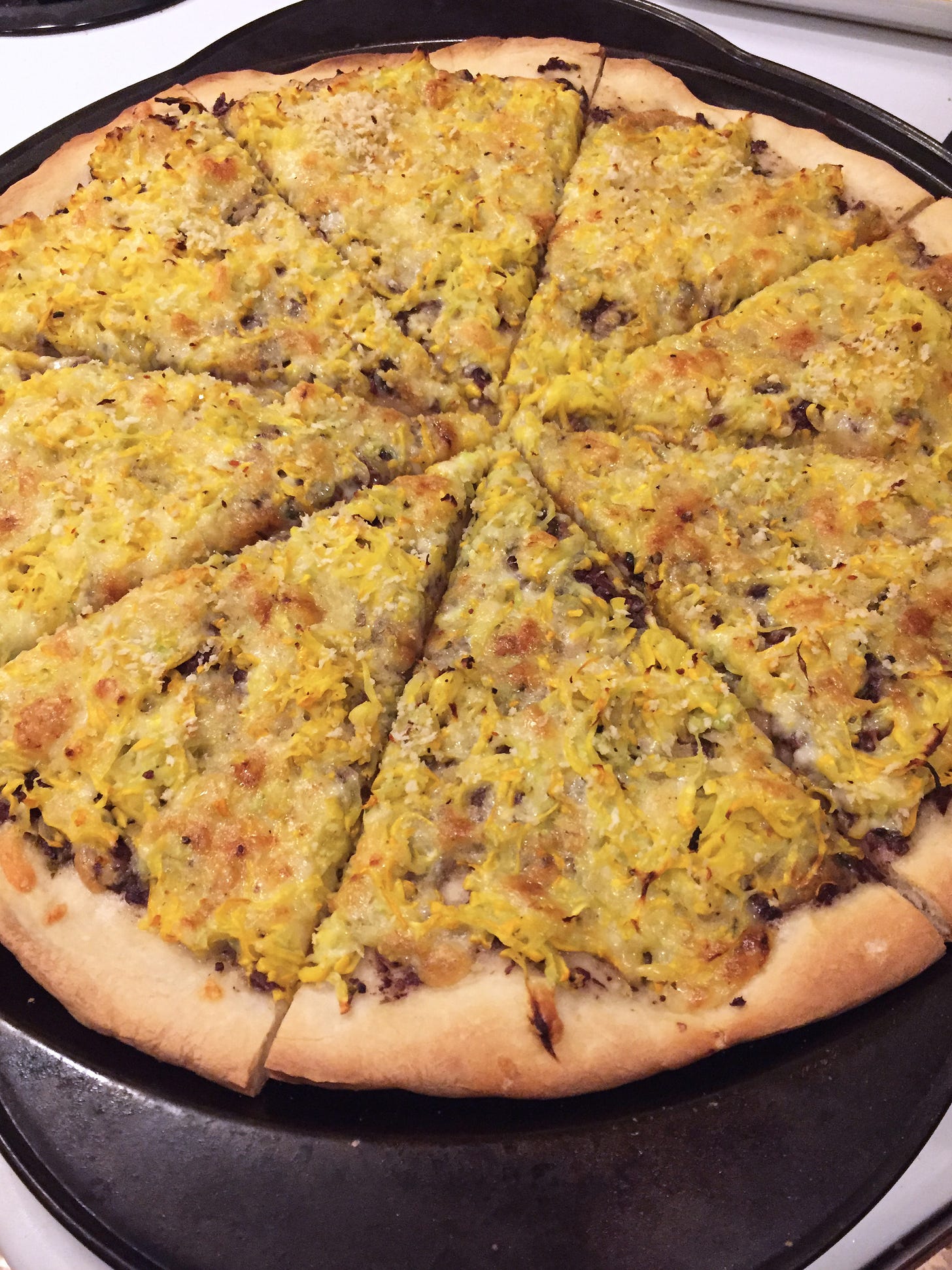 a large pizza sliced into pieces sits in its pan on the stove. Browned cheese and shreds of yellow zucchini cover the top, and bits of dark tapenade are visible in spots underneath, and at the edges.