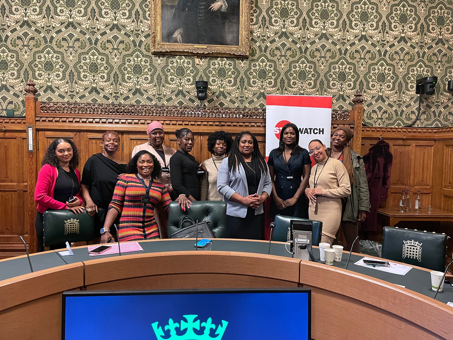 Women standing behind a table in an ornately decorated room in parliament, with Bell Ribeiro-Addy, all smiling at the camera