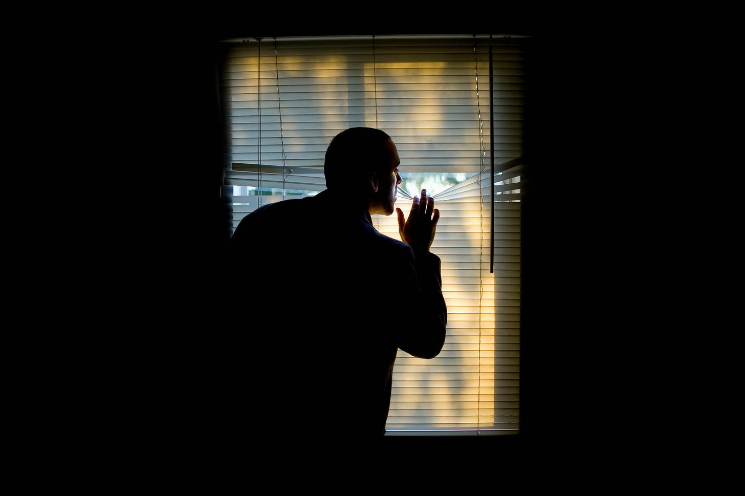 Man in a dark room peering out  through levolor blinds