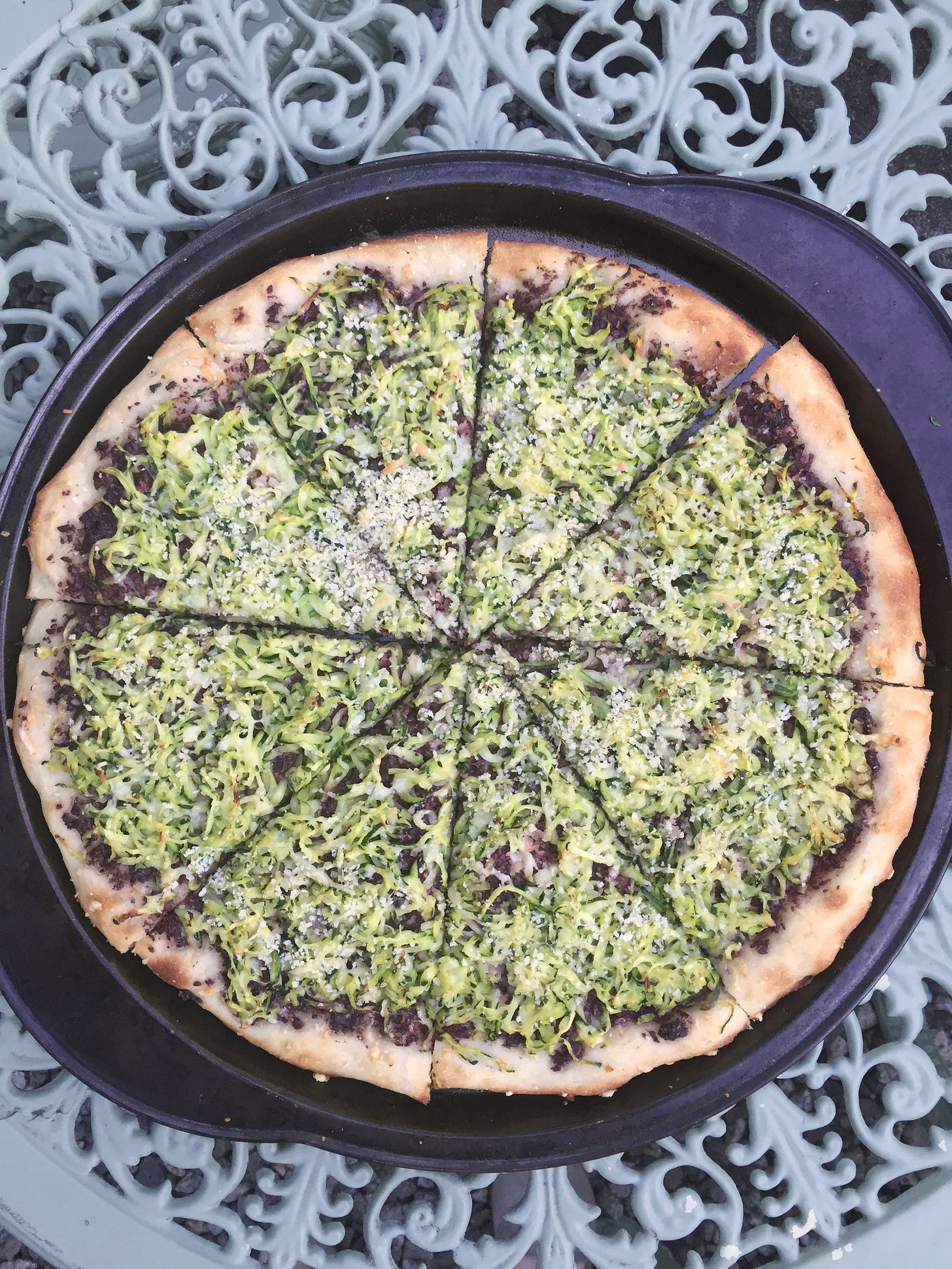 on a blue-green wrought iron table, a large pizza pan containing a pizza with shredded zucchini, breadcrumbs, and black olive tapenade. The crust is browned and bubbled at the edges.