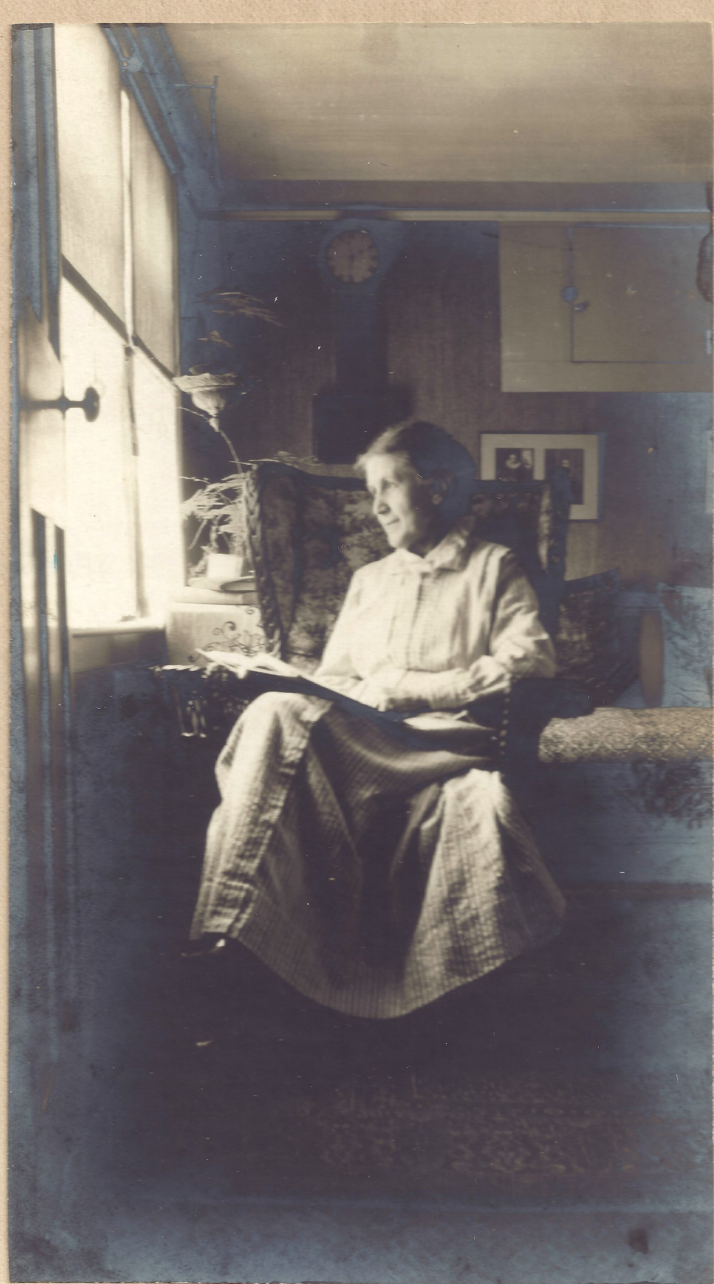 black and white photo of woman in her mid-60s, wearing long skirt and frilled blouse, gazing out window with notebook in her lap. 