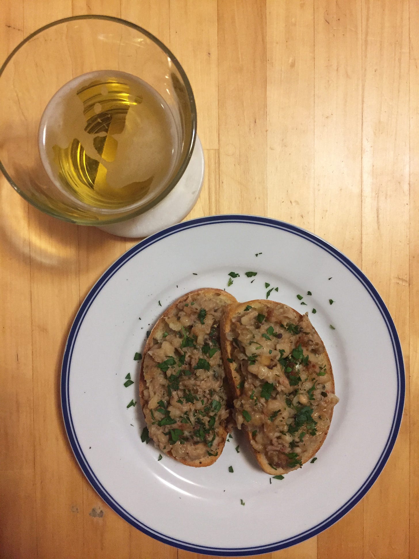On a white plate with a blue rim are two pieces of toast with the bean and tuna casserole on top, scattered with parsley. Next to the plate is a pale glass of beer.