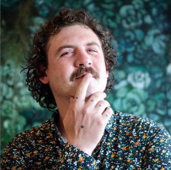 A picture of Zak Spence is here again an emerald forest background. He is a white man with blue eyes and curly light brown hair. He has a mustache and a sterling septum piercing. He is bringing his hands up to his face, and the black dot tattoos on the back of his hand and each of his knuckles is visible.