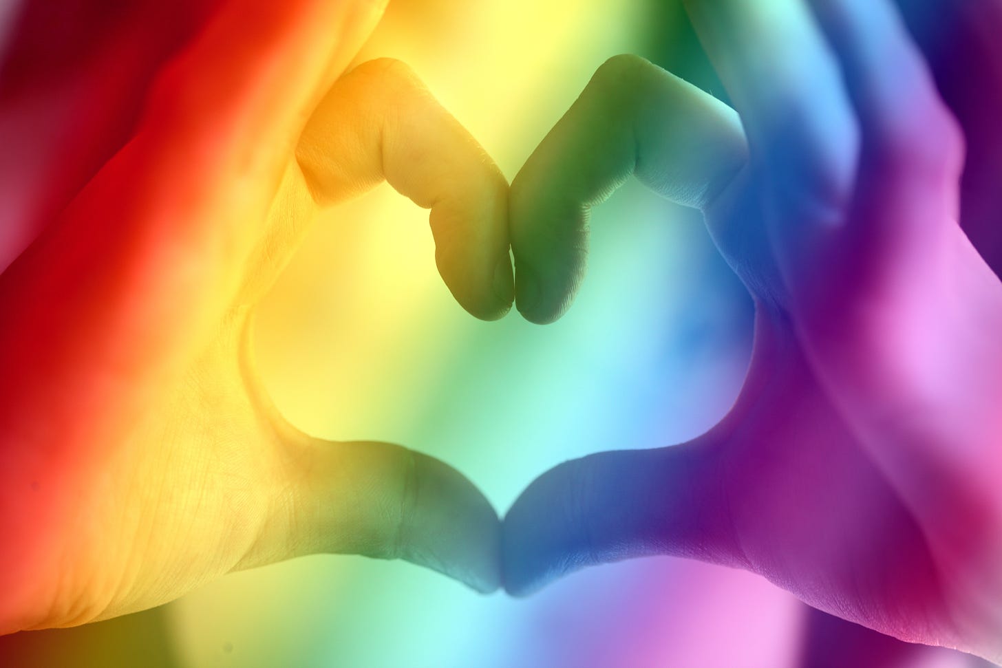 A woman makes a heart shape with her hands as rainbow light reflects on them in a diagonal pattern from the upper left hand corner to the lower righthand corner from dark red to light purple. I think they're white hands but it's hard to tell. It's a real artsy fartsy photo 