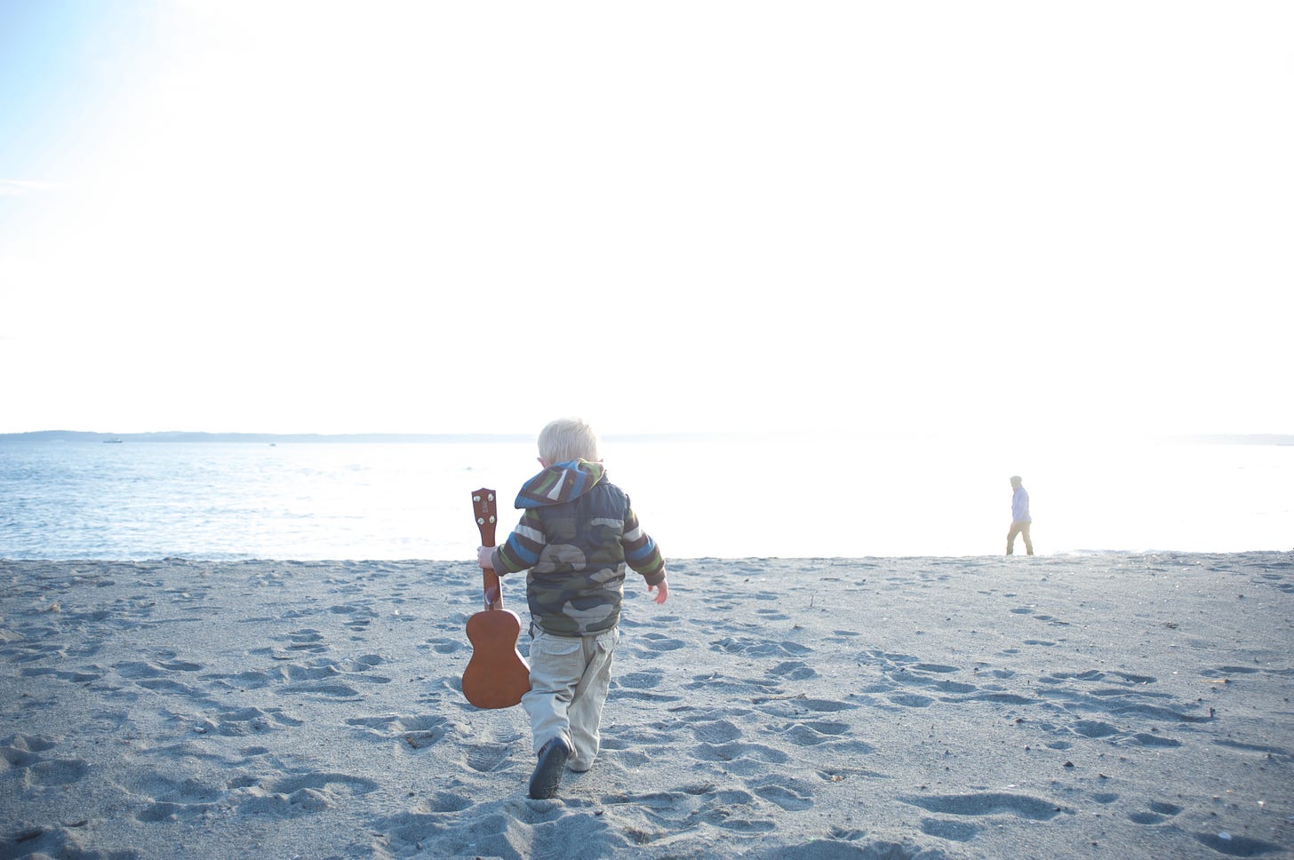Child in a camouflage coat carrying a toy guitar on a beach