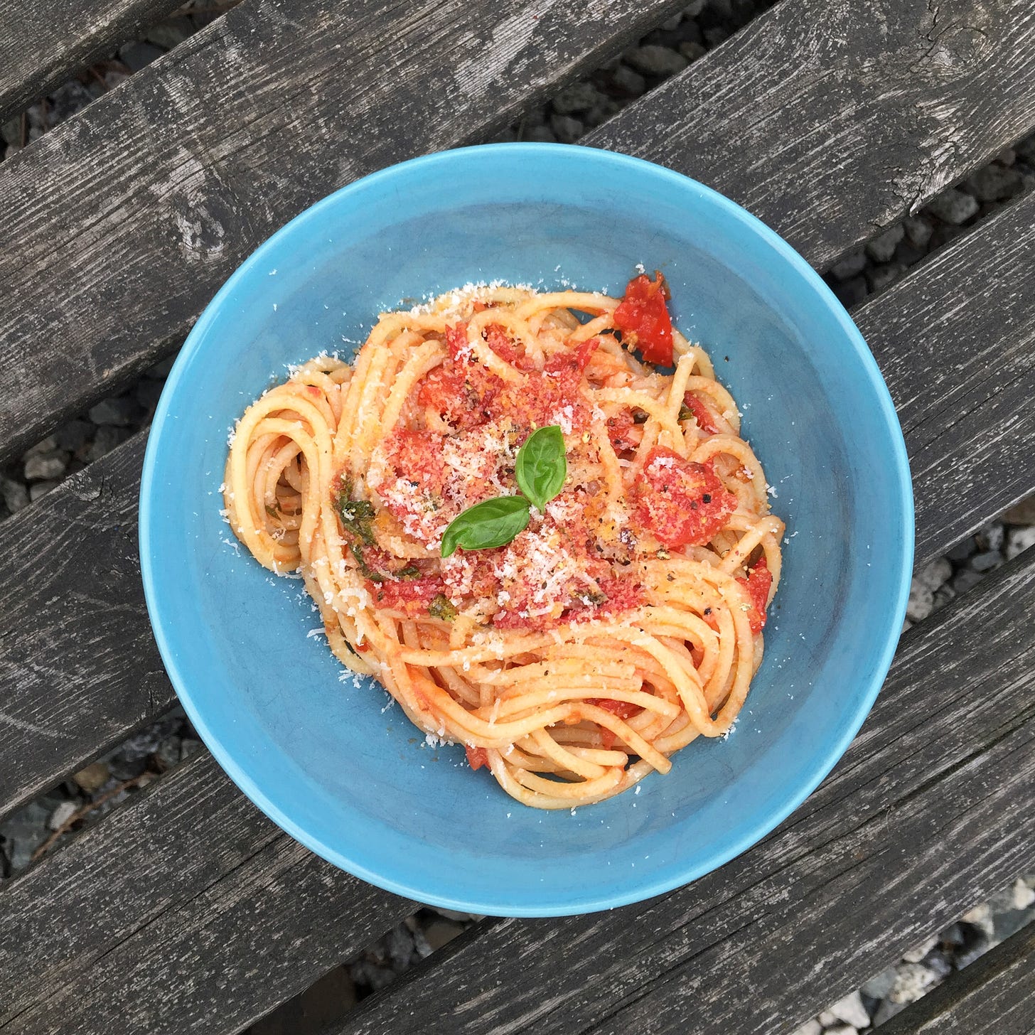 On a wooden bench, a blue bowl of spaghetti with pomodoro sauce, with parmesan and two small basil leaves on top.