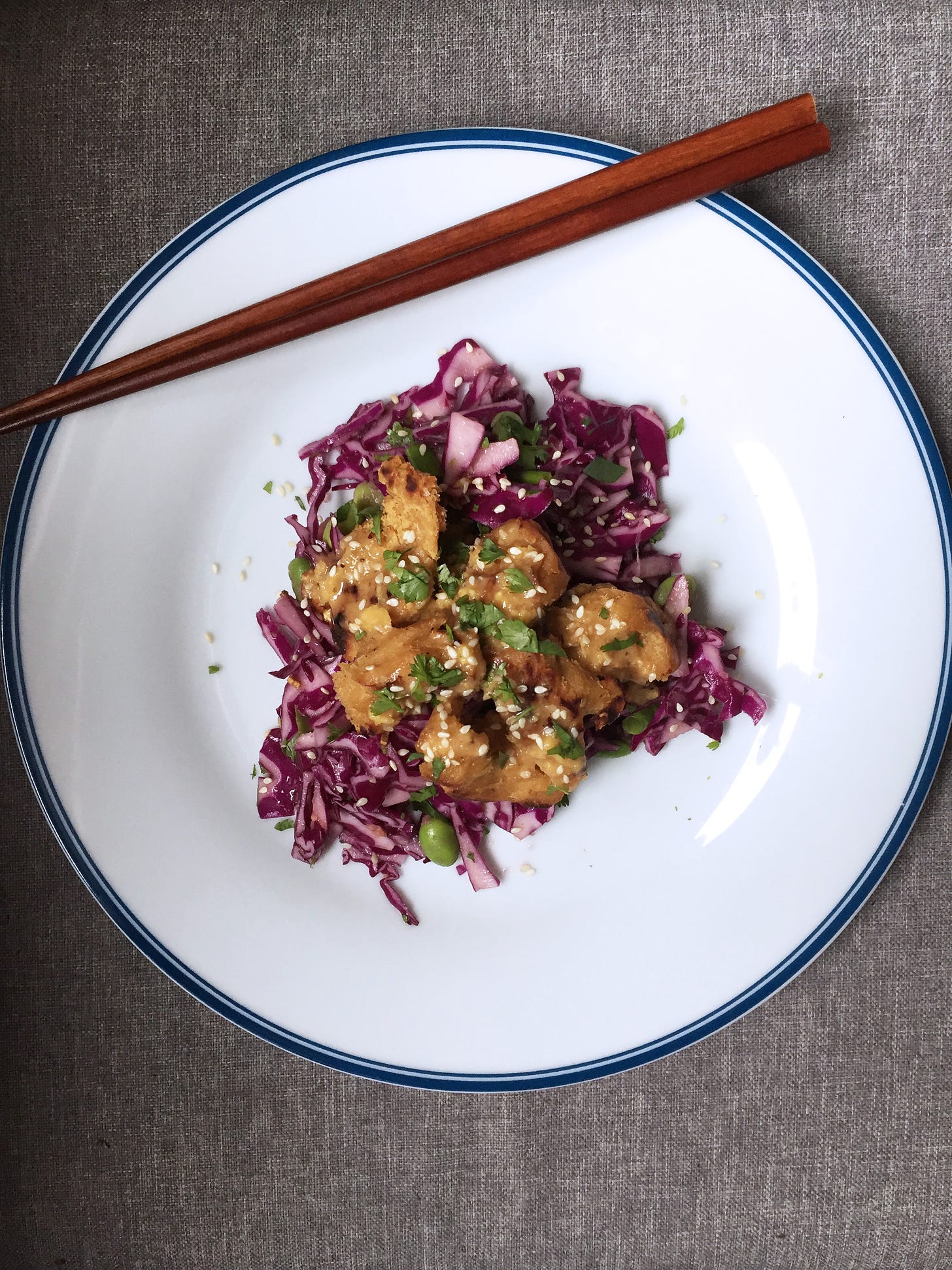 A white plate from above with brown chopsticks in the upper corner. In the center is a red cabbage slaw with edamame visible, and seitan, sesame seeds, and cilantro on top.