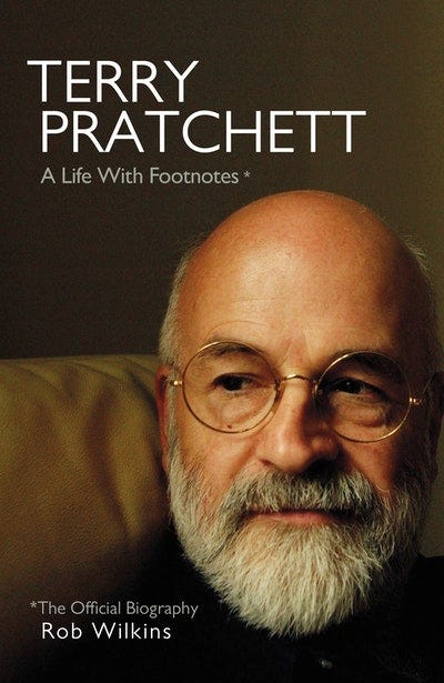 A photo of a bald, older, male-presenting person with a grey beard, glasses, and a crooked nose. Over the image are the words ‘Terry Pratchett: A Life with Footnotes* *The Official Biography, Rob Wilkins’.
